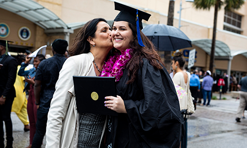 student standing on graduation day with cap and gown on and their mother embracing them