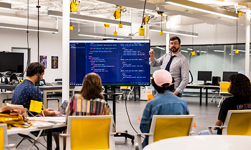 professor stands in a computer lab teaching a small group of students