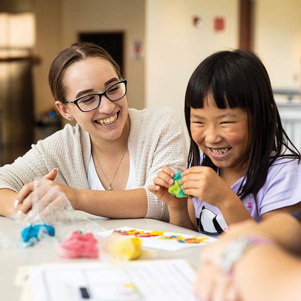 Teaching student sitting next to an elementary aged student playing with clay
