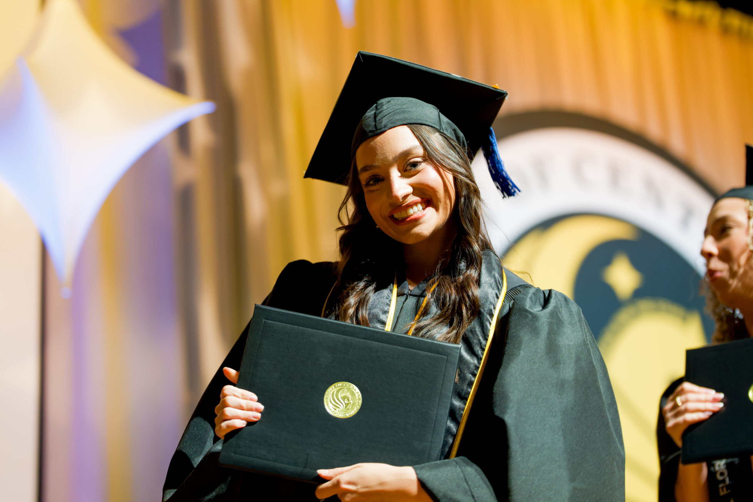 UCF graduate smiles holding diploma on the graduation stage